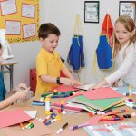 Three children sit around a table in a classroom, engaged in arts and crafts with Medium Weight Construction Paper, glue sticks, and markers. Classroom walls are decorated with colorful posters and aprons hang in the background.