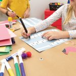 Children crafting with Medium Weight Construction Paper, markers, and glue sticks at a table. One child is drawing on a blue-striped sheet while another reaches for supplies. Various craft materials are scattered around.