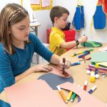 Two children are crafting at a table using Medium Weight Construction Paper, glue sticks, markers, and other art supplies. The background shows colorful storage units and aprons hanging on hooks.