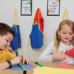 Two children engage in arts and crafts, one cutting red Medium Weight Construction Paper with scissors and the other drawing on purple Medium Weight Construction Paper with a marker. Colorful aprons and encouraging posters are visible in the background.