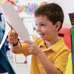 A young boy in a yellow shirt is smiling while cutting a piece of Medium Weight Construction Paper with scissors in a classroom setting.