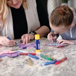 Two people are crafting at a table, using markers and glue to decorate cutouts made from Medium Weight Construction Paper. Assorted craft supplies are scattered on the surface.