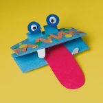 A vibrant paper craft monster made using Construction Paper Samplers, featuring a blue face, googly eyes, a red tongue, and decorative patterns, set against a bright yellow background.