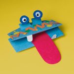 A playful monster craft created from Medium Weight Construction Paper in blue, featuring googly eyes, vibrant spots, and a long red tongue, all set against a bright yellow background.